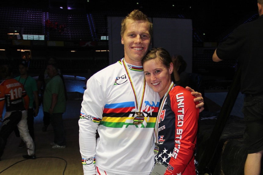 Sam stands with his arm around Alise while wearing a gold medal and a rainbow jersey