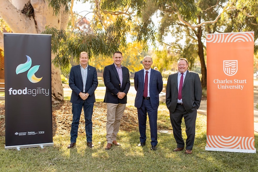 Four men in suits between two signs in an outside area. 