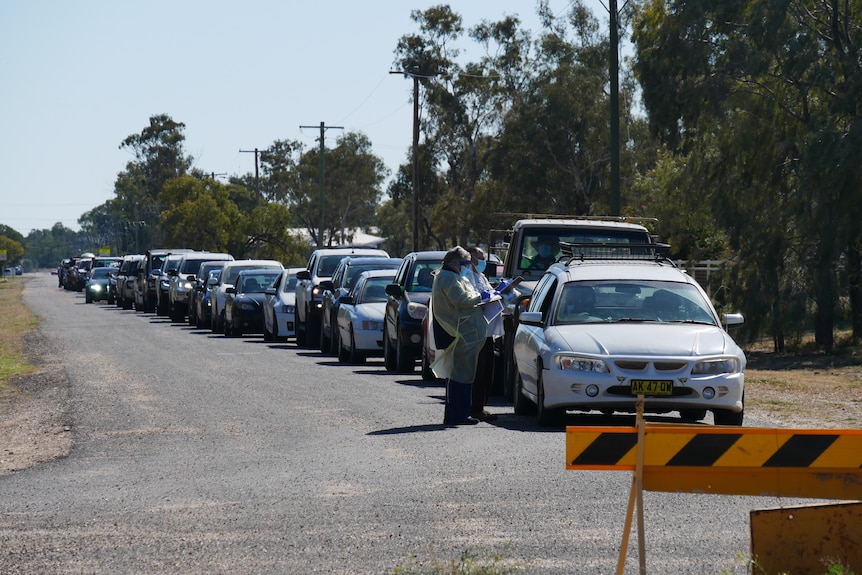 Two health workers in full PPE standing beside the first of a long line of vehicles. They are carrying clipboards 