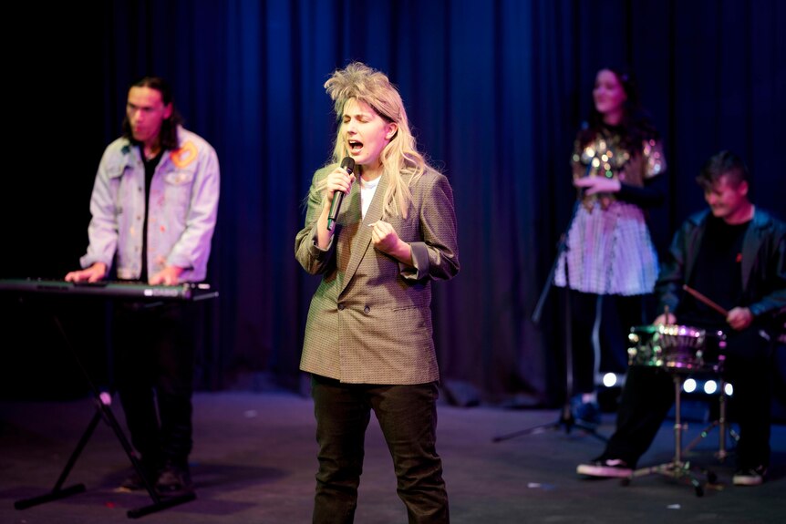 A teenage girl wearing a blonde '80s mullet and brown blazer on stage singing a note with her eyes closed