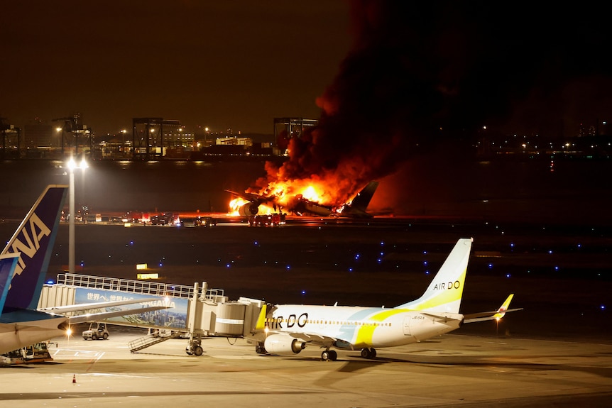 Bright orange flames engulf a plane on the tarmac at an airport.