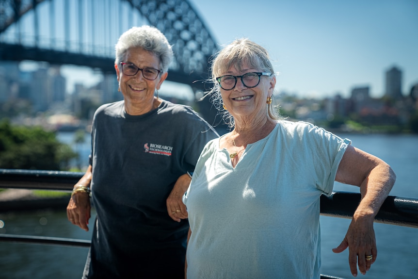 Two women stand in front of the Sydney Harbour Bridge on a sunny day. 