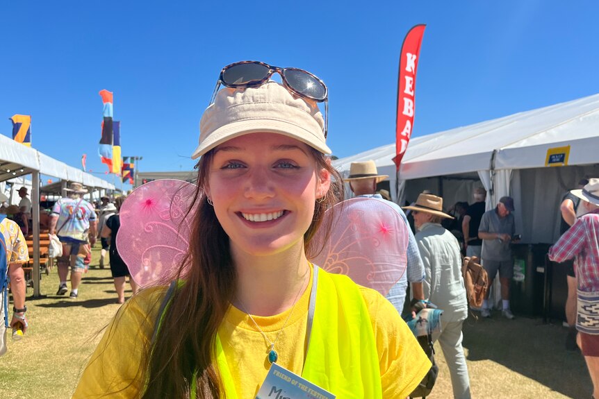A young woman at a festival wearing a yellow shirt, pink fairy wings, and a white baseball cap with sunglasses perched on top.