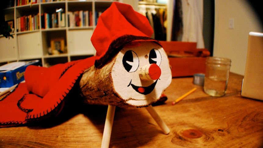 A small log with a smiling face painted on and two front legs wears a red woollen cap and red blanket.