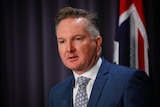 Chris Bowen in a navy suit and tie in front of the Australian flag at a press conference.