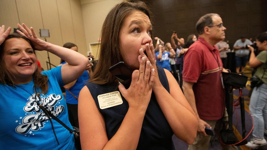 A woman with long dark hair in a black shift dress puts her hands her over mouth in a crowded room 