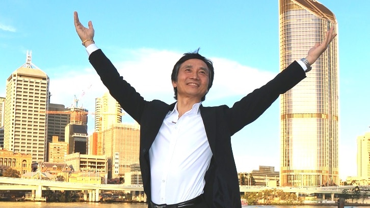 Li Cunxin striking a ballet pose at Brisbane's South Bank, with river and CBD in background.