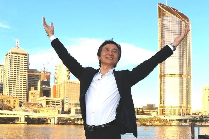 Li Cunxin striking a ballet pose at Brisbane's South Bank, with river and CBD in background.