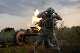A soldier in protective camouflage gear ducks and holds his helmet as a mortar explodes into the sky behind him