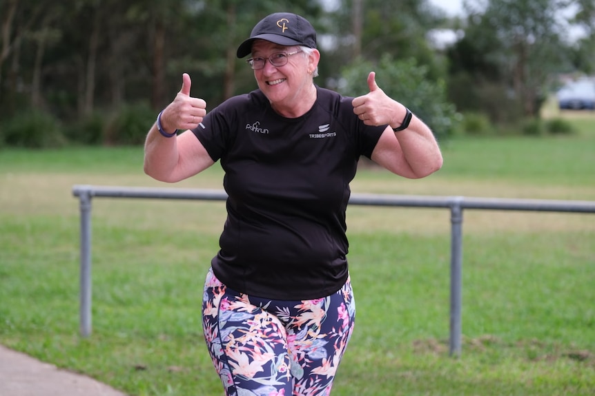 A woman running puts both thumbs up and smiles at the camera