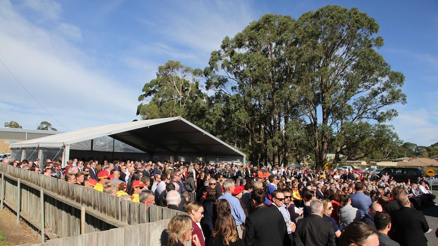 A massive crowd gathers in a car park for a funeral in a country town.