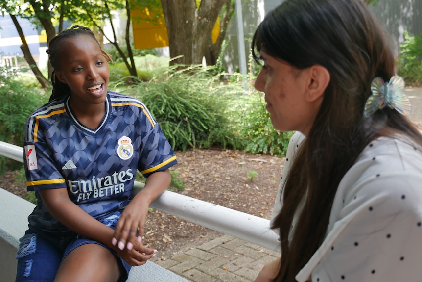 Two girls are pictured talking to each other, one is wearing a football shirt, the other a blouse with polka dots.