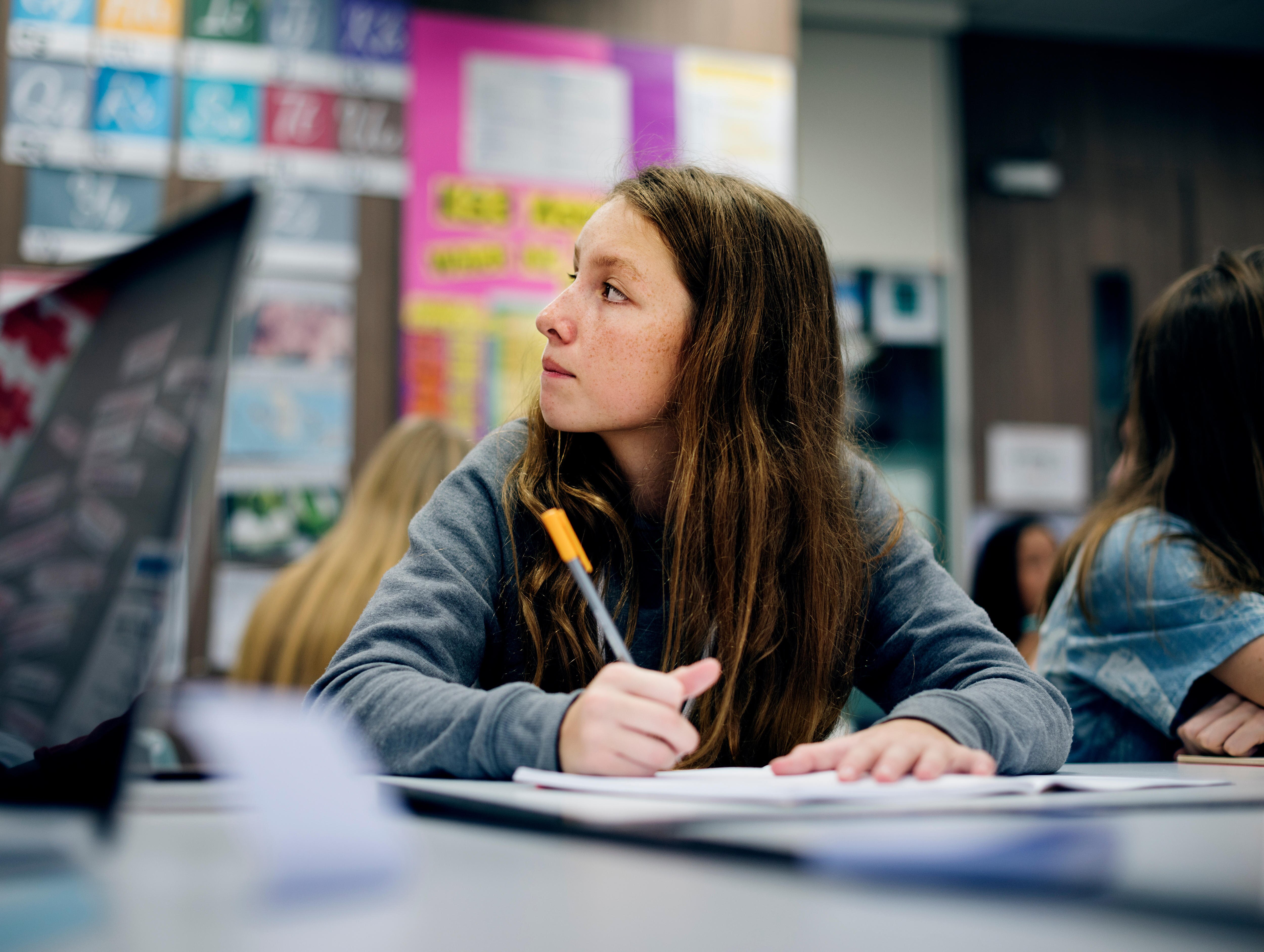 A young female student looks to the left of the classroom while working at her desk.