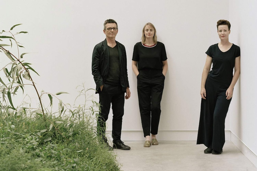 The three creatives stand against a white wall, wearing black, with their botanical installation peaking into the frame.