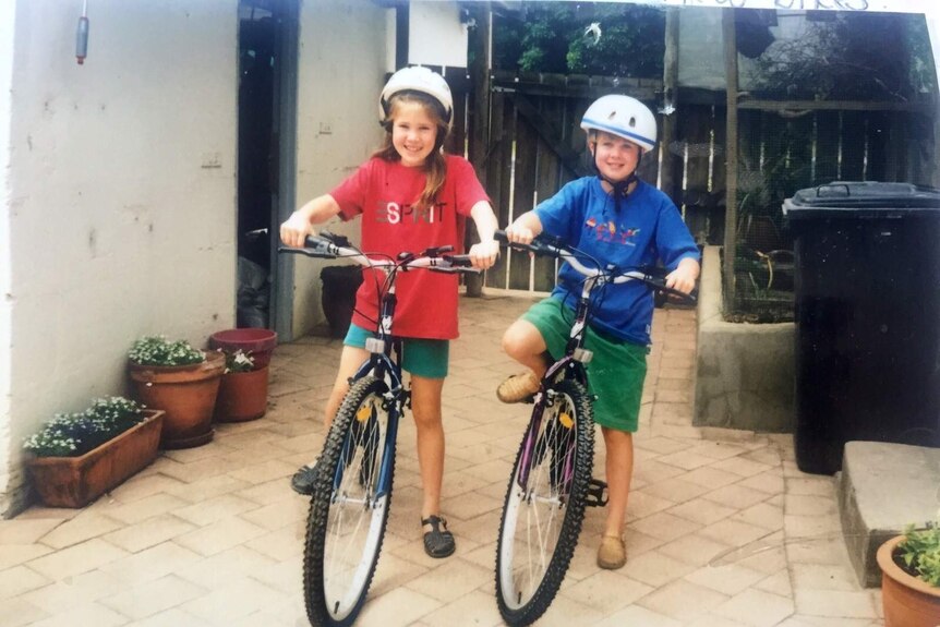 Jess and her sister are standing smiling on bikes in a backyard, wearing helmets.