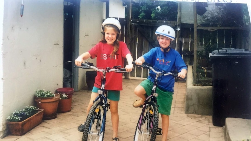 Jess and her sister are standing smiling on bikes in a backyard, wearing helmets.