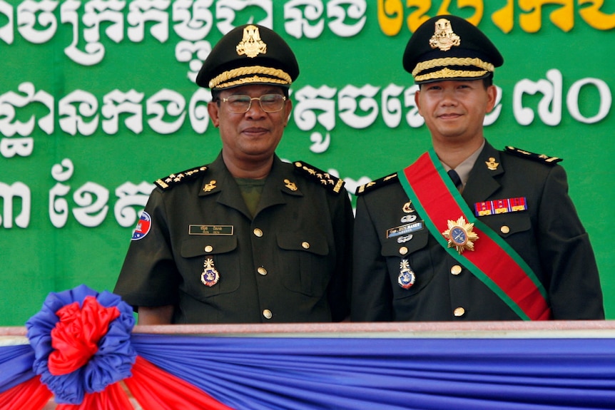 Two men in formal military uniform, dark green with gold details and gold-trimmed hats, stand side by side on stage