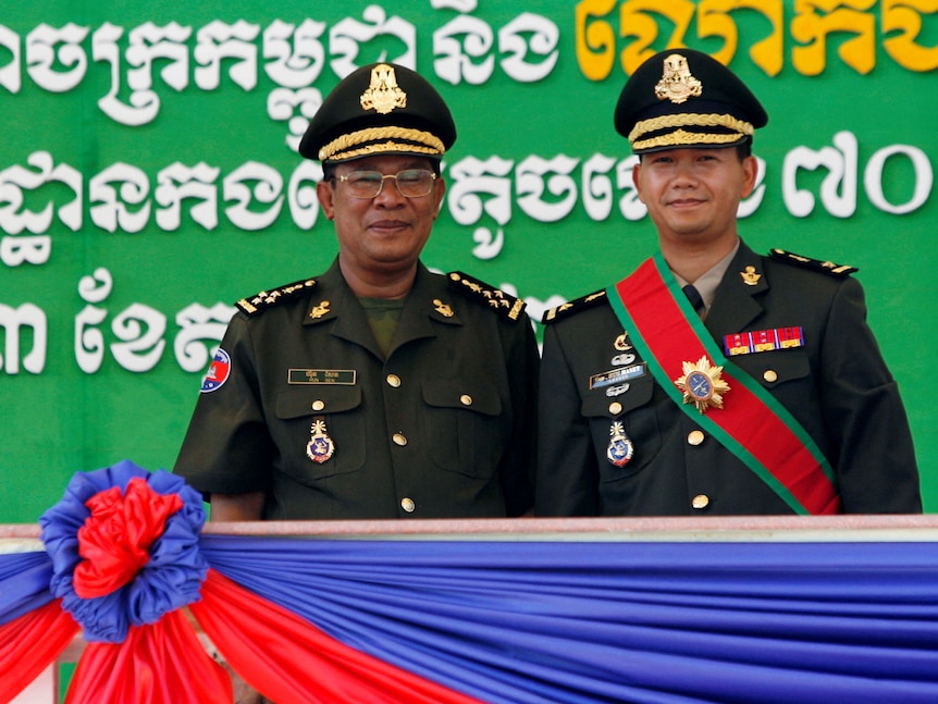 Two men in formal military uniform, dark green with gold details and gold-trimmed hats, stand side by side on stage