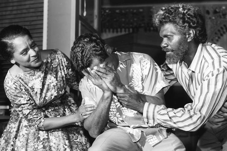 Black and white photo of a woman and man leaning in to support man between them, who sits with his face in his hands.