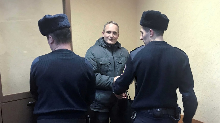 A man in a khaki thick jacket is seen between two Russian police officers with their backs to the camera, one opening a door.