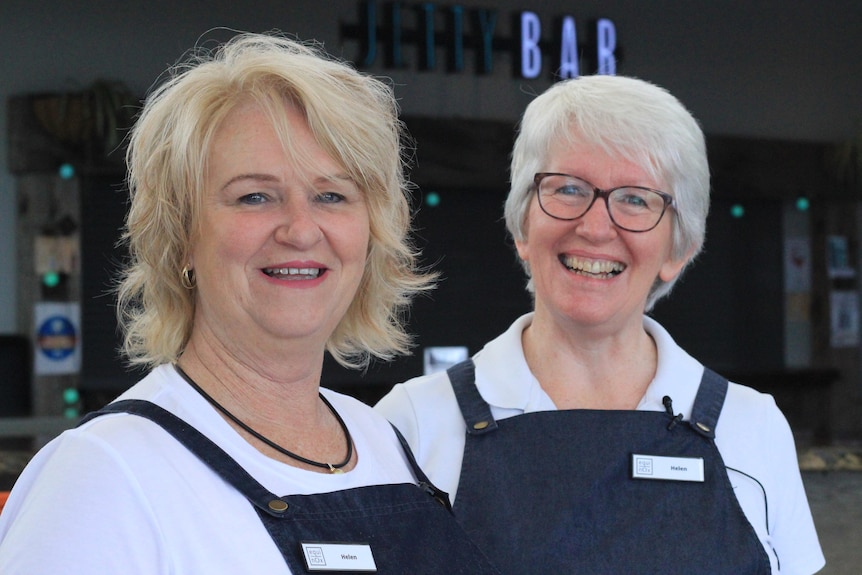 Two women, in white shirts and work aprons, smiling in front of a "Jetty Bar" sign. 