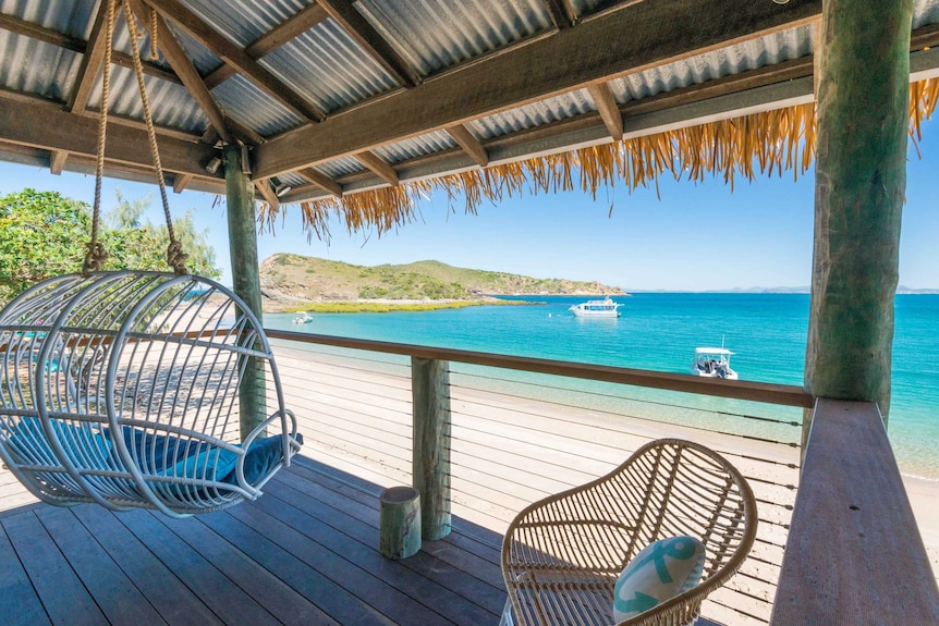 Looking out from a timber deck with thatched roof and swing chair looking out over blue water, boats, green hills and white sand