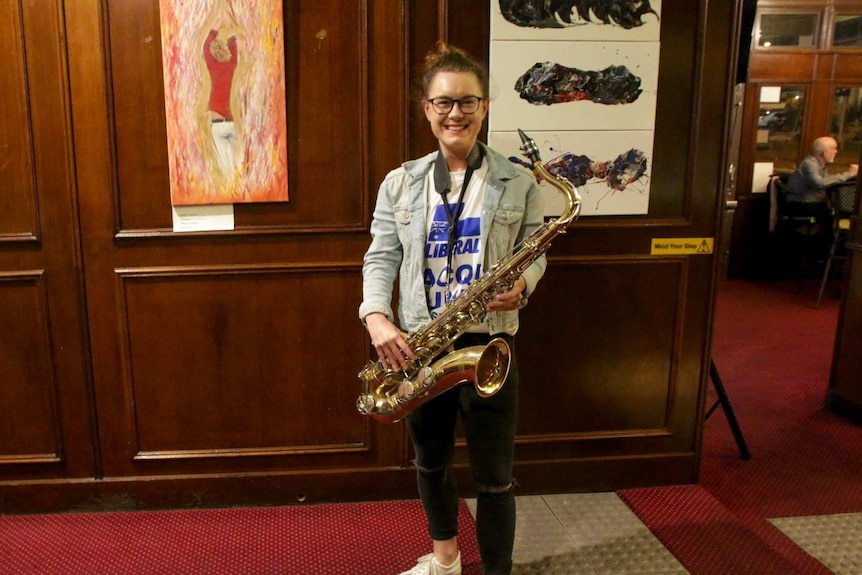 Jacqui Munro, a young Liberal candidate at the 2019 federal election, stands in a pub holding her saxophone.