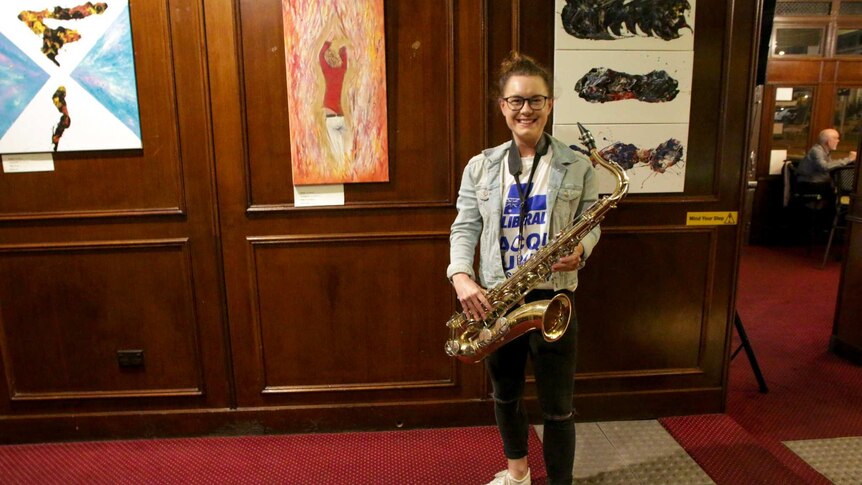 Jacqui Munro, a young Liberal candidate at the 2019 federal election, stands in a pub holding her saxophone.