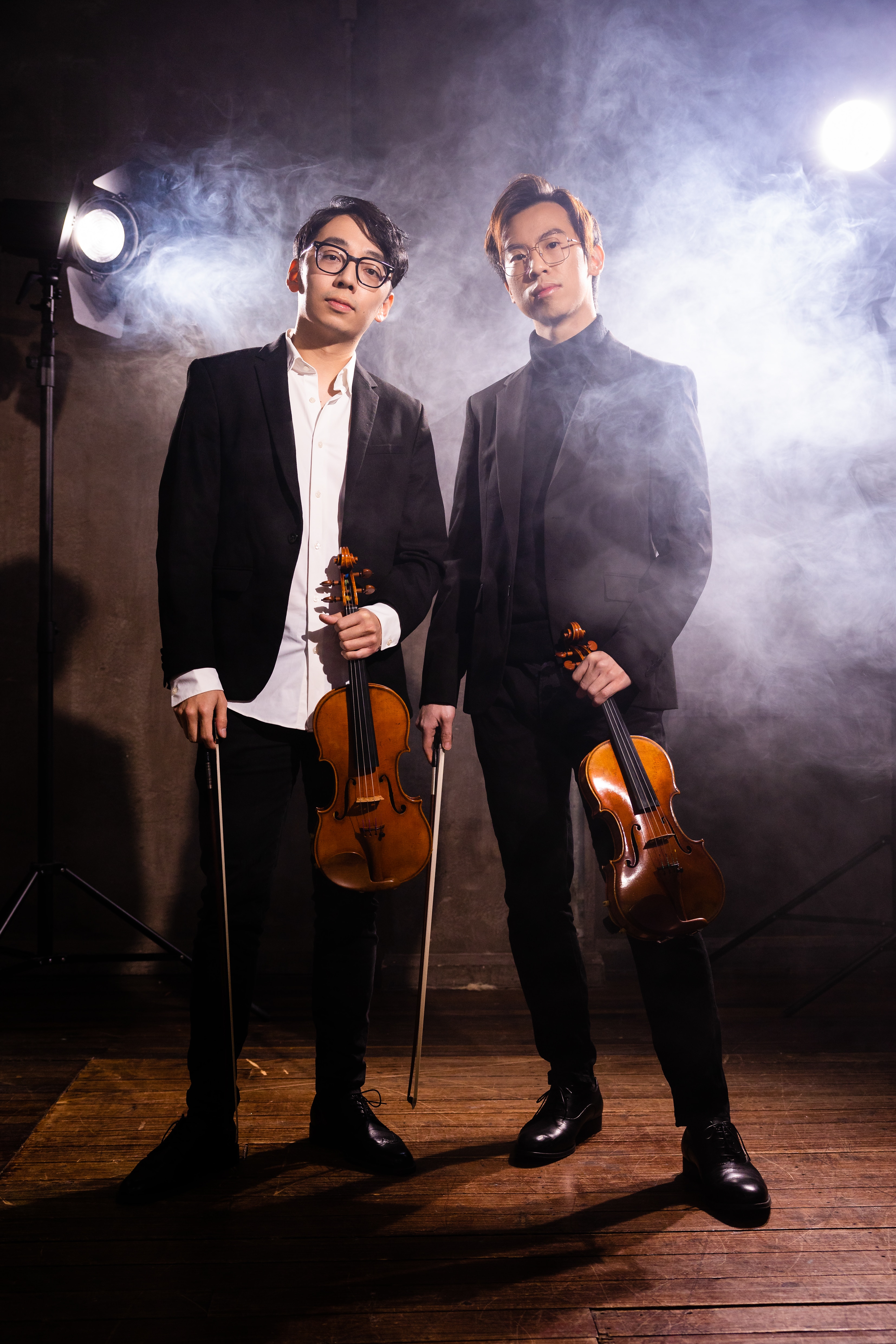 Brett and Eddy wearing suits, looking serious, smoke behind them, holding violins.