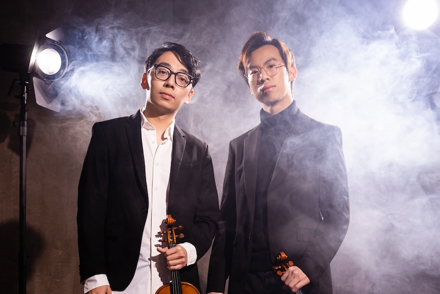 Brett and Eddy wearing suits, looking serious, smoke behind them, holding violins.