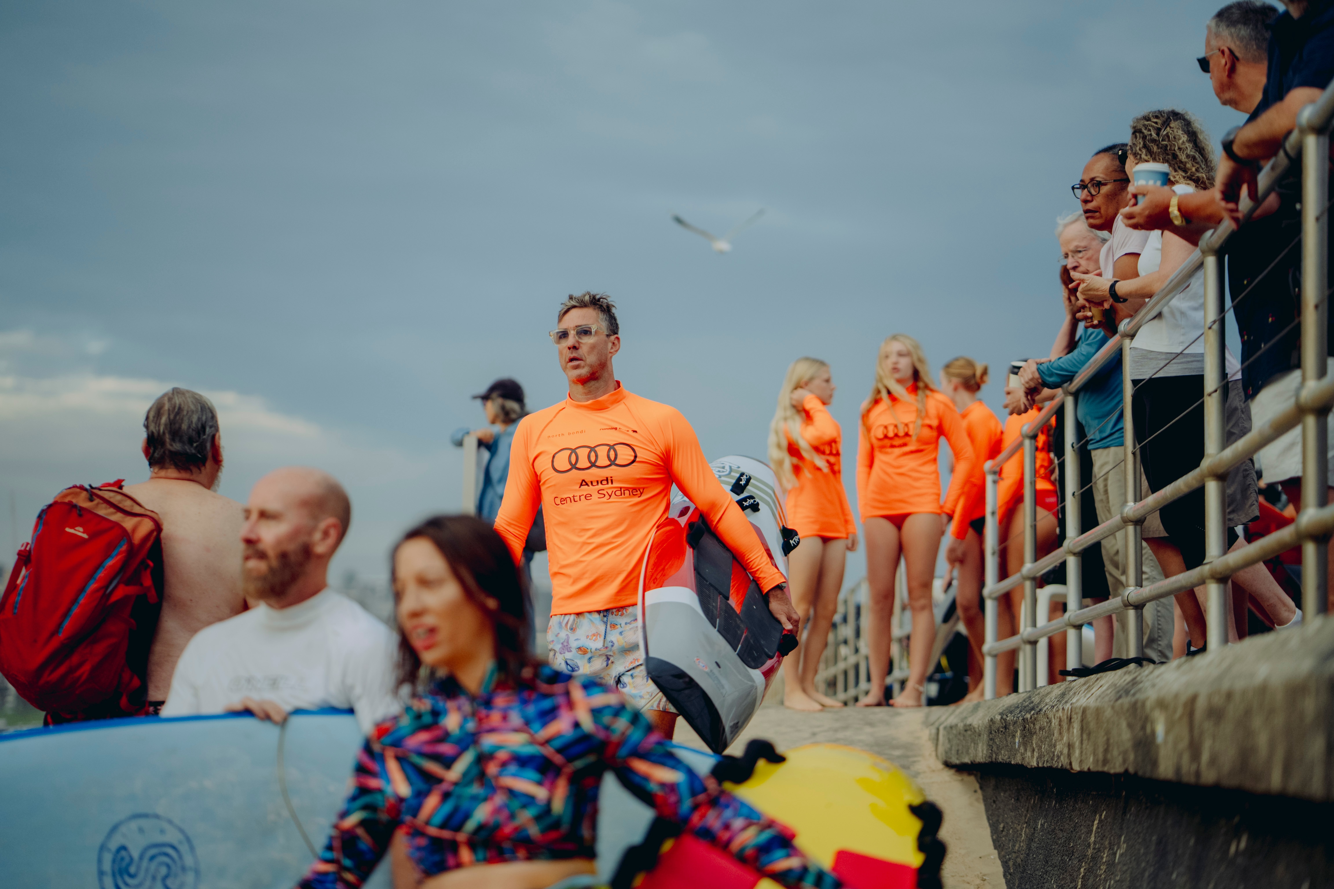 Um homem com uma prancha de surf desce as escadas até a praia enquanto pessoas apoiadas em uma grade assistem ao evento.
