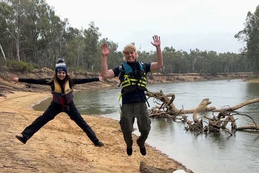 A young woman and teenage boy wearing life jackets jumping for joy on a river bank.