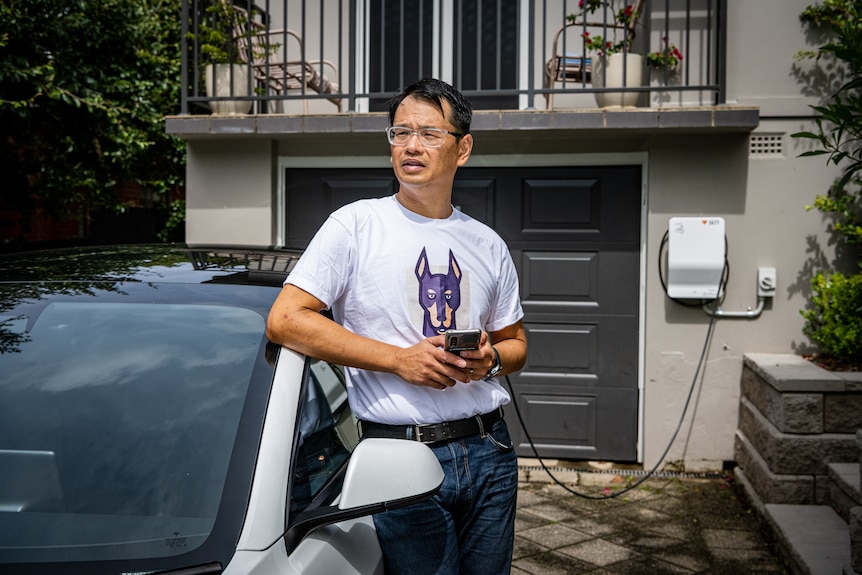 Marc Tan leans against a Tesla EV that's plugged into a smart charger outside his home, holding a mobile phone.
