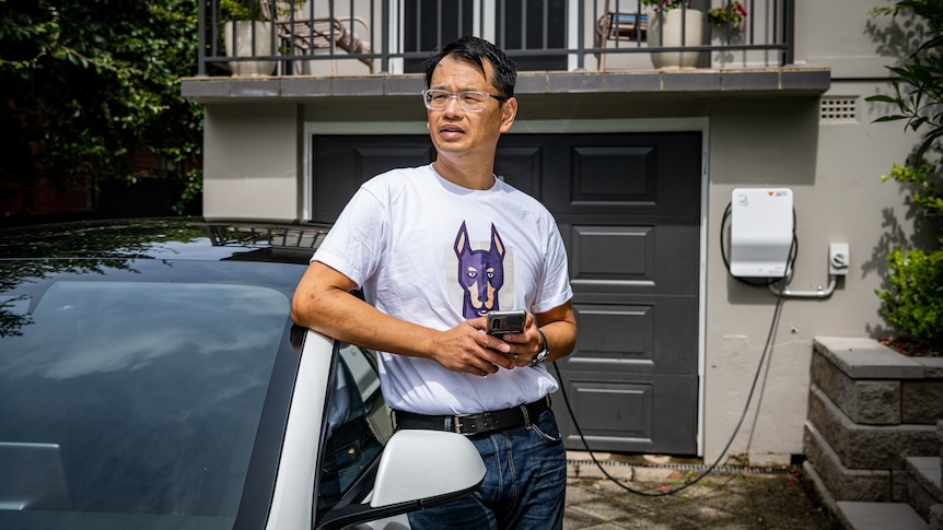 Marc Tan leans against a Tesla EV that's plugged into a smart charger outside his home, holding a mobile phone.