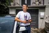 Marc Tan leans against a Tesla EV that's plugged into a smart charger outside his home, holding a mobile phone.