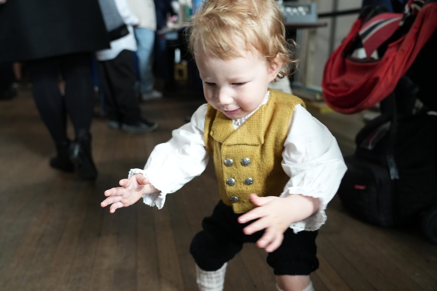 A toddler boy in traditional Norwegial dress holds his hands out and tries to balance.