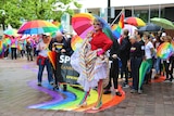 People dressed in bright rainbow colours stand on a rainbow mural in Canberra.