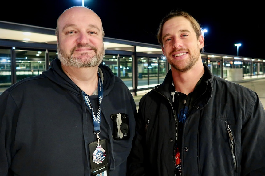 two men at night stood near a bus station smiling at camera