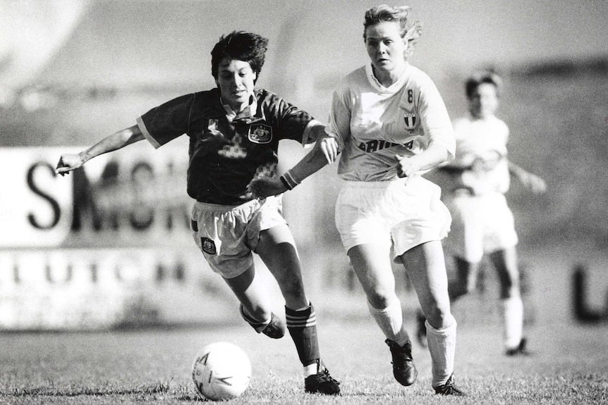 Moya Dodd, left, runs while looking down at a ball while pushing past a woman wearing a light-coloured top