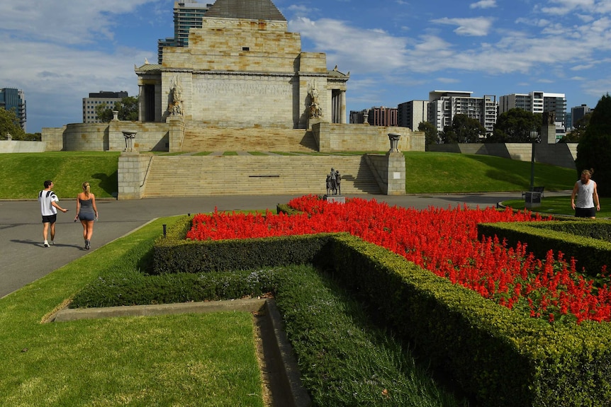 Melbourne's shrine of remembrance is almost abandoned. a few people walk around a cross-shaped hedge filled with red flowers