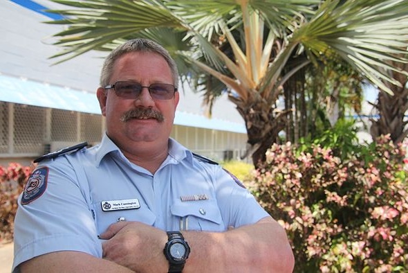 A man in an NT Police uniform stands outside, in front of fan palms, with his arms crossed over his chest.