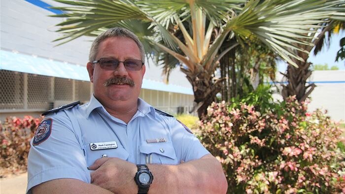 A man in an NT Police uniform stands outside, in front of fan palms, with his arms crossed over his chest.