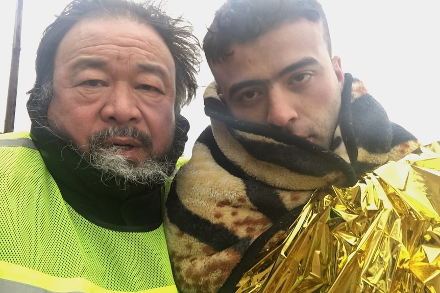 Artist Ai Weiwei standing next to a refugee man wrapped in a thermal blanket
