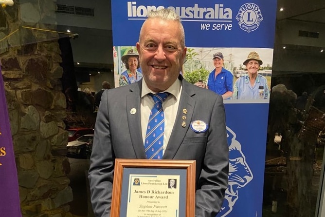 An elderly man with white hair wearing a grey suit and blue tie holds up an award for his contribution with the Lions Australia.