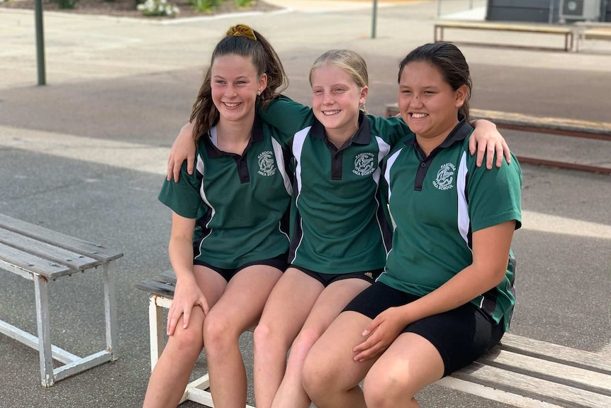 Three young female girls sitting on a wooden bench with their arms wrapped around each other.