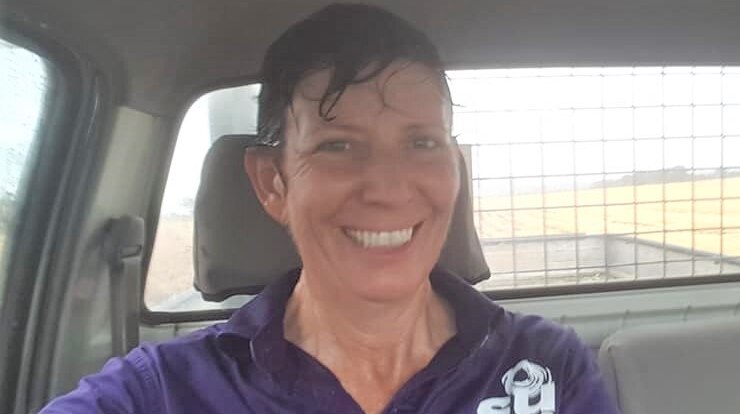 A woman with wet hair and tshirt sits smiling in a car while it rains outside