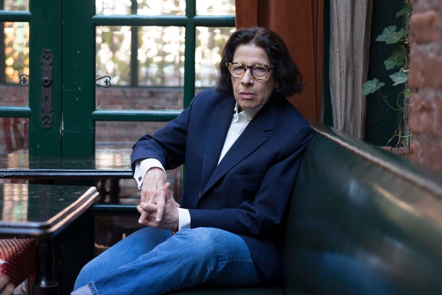 Fran Lebowitz sits on a greenleather bench in a navy blazer, white shirt and blue jeans. She is looking directly at the camera.