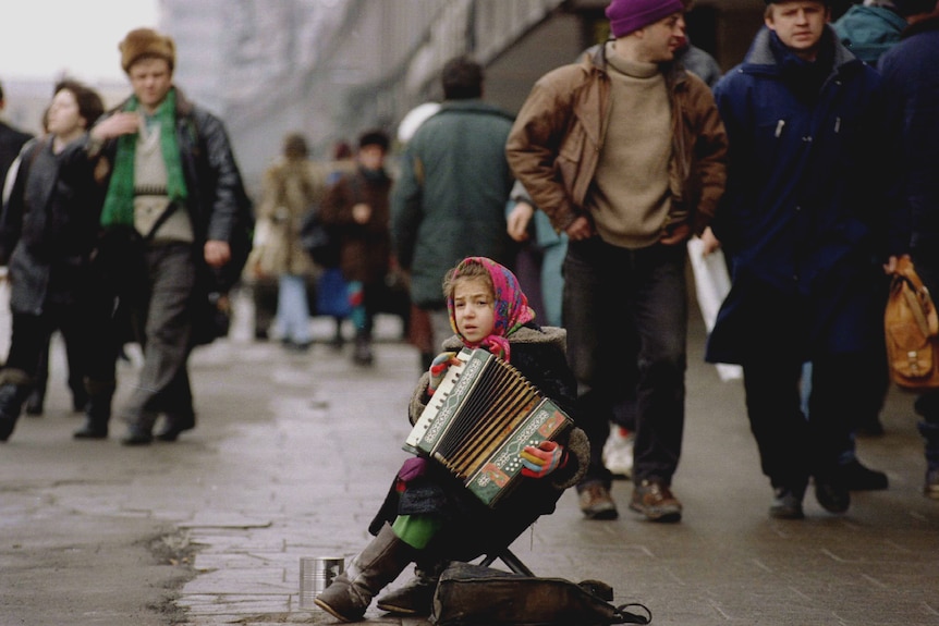 A young girl sits in a street playing an accordian as people walk by dressed in coats.