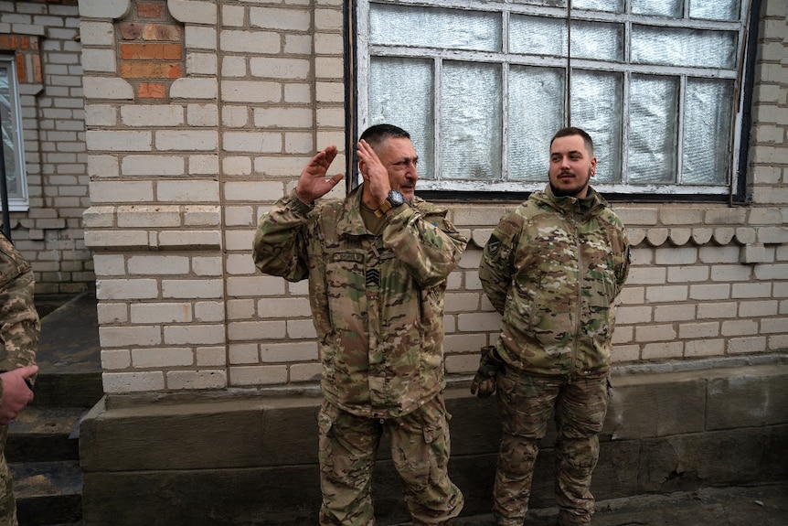 A man in military uniform gestures with his hands while another man, also in uniform,  looks on