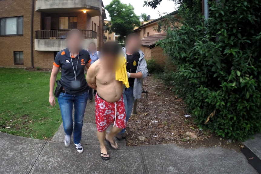 A shirtless man is led from a unit block by police officers.